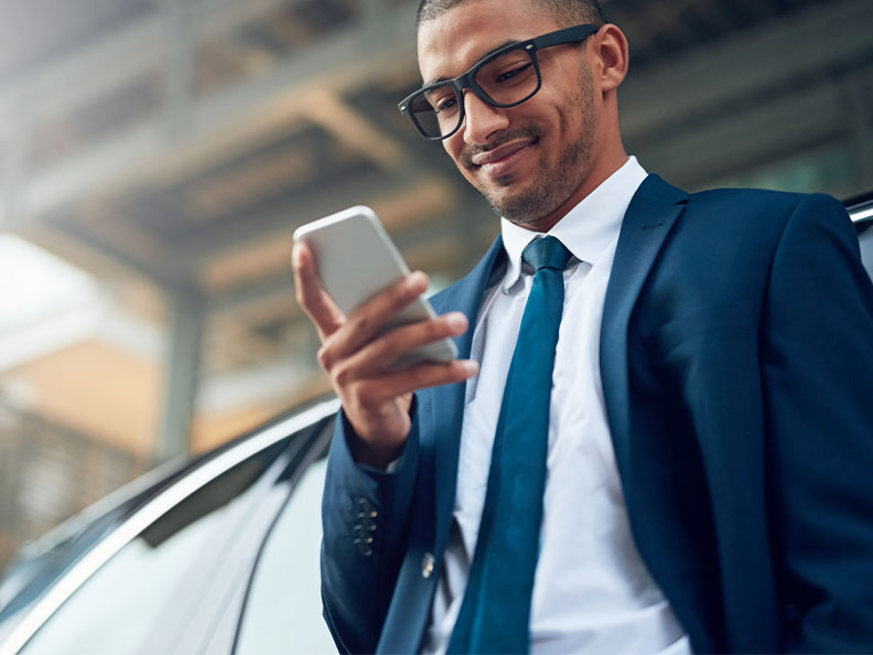 Shot of a young businessman using his phone while standing outside leaning against a car. He’s wearing a suit.