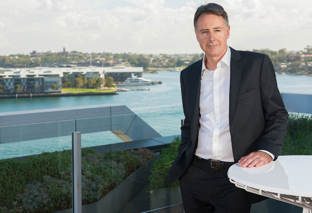 A business man in a suit standing outside on an office rooftop with a hand on a table. Macquarie employee - David Barrett