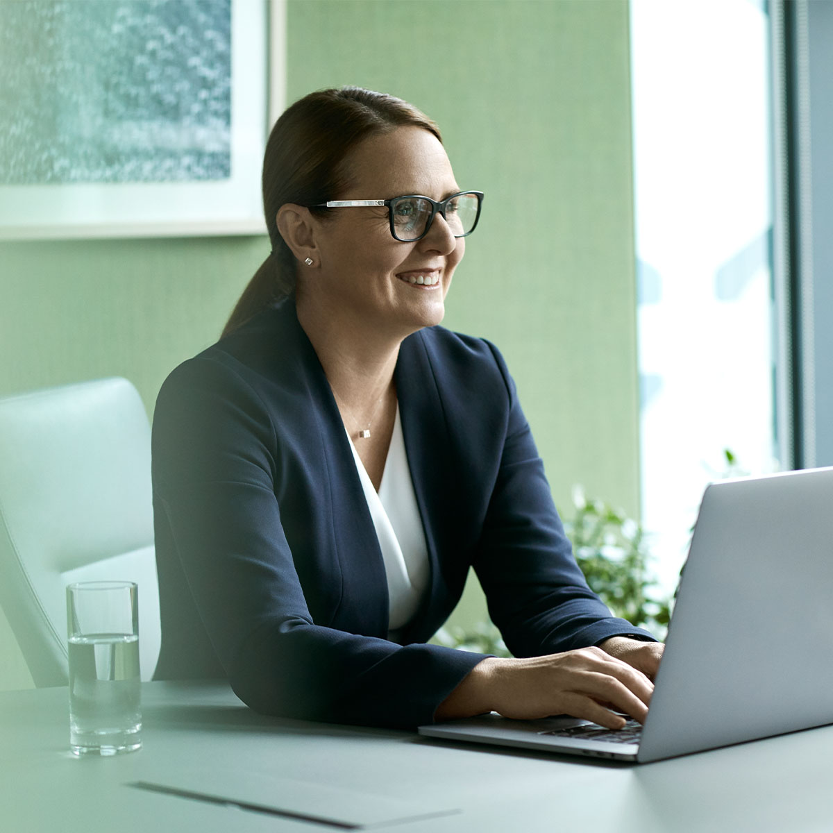 A businesswoman with glasses sitting at a desk working on laptop and smiling