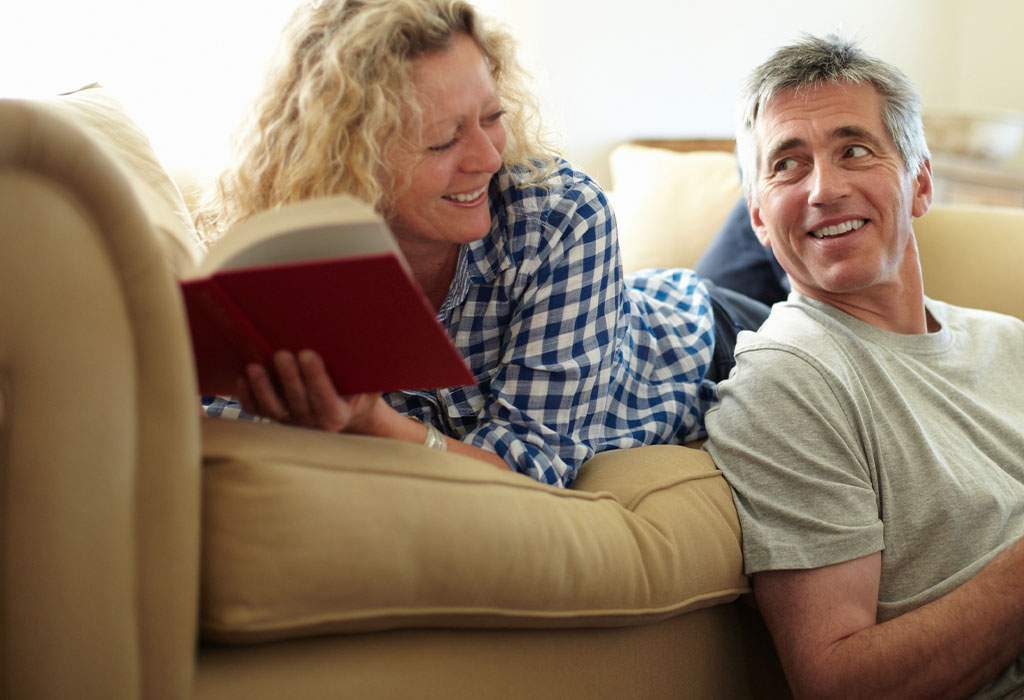 A couple looking at eachother as they read books together in their lounge room on their couch in their home. A man is using a tablet and a woman is holding a hardcover book.