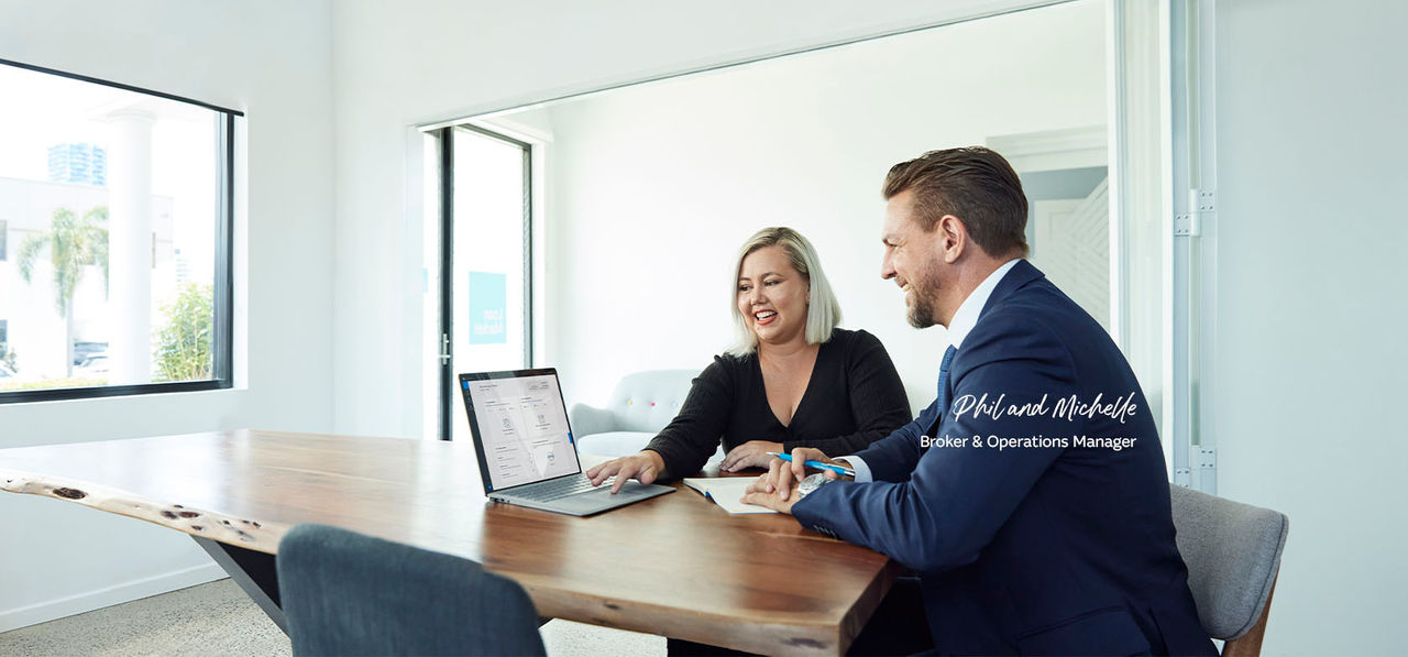 Phil and Michelle Loan Market Edge sitting in white office at wood table smiling with broker portal on laptop