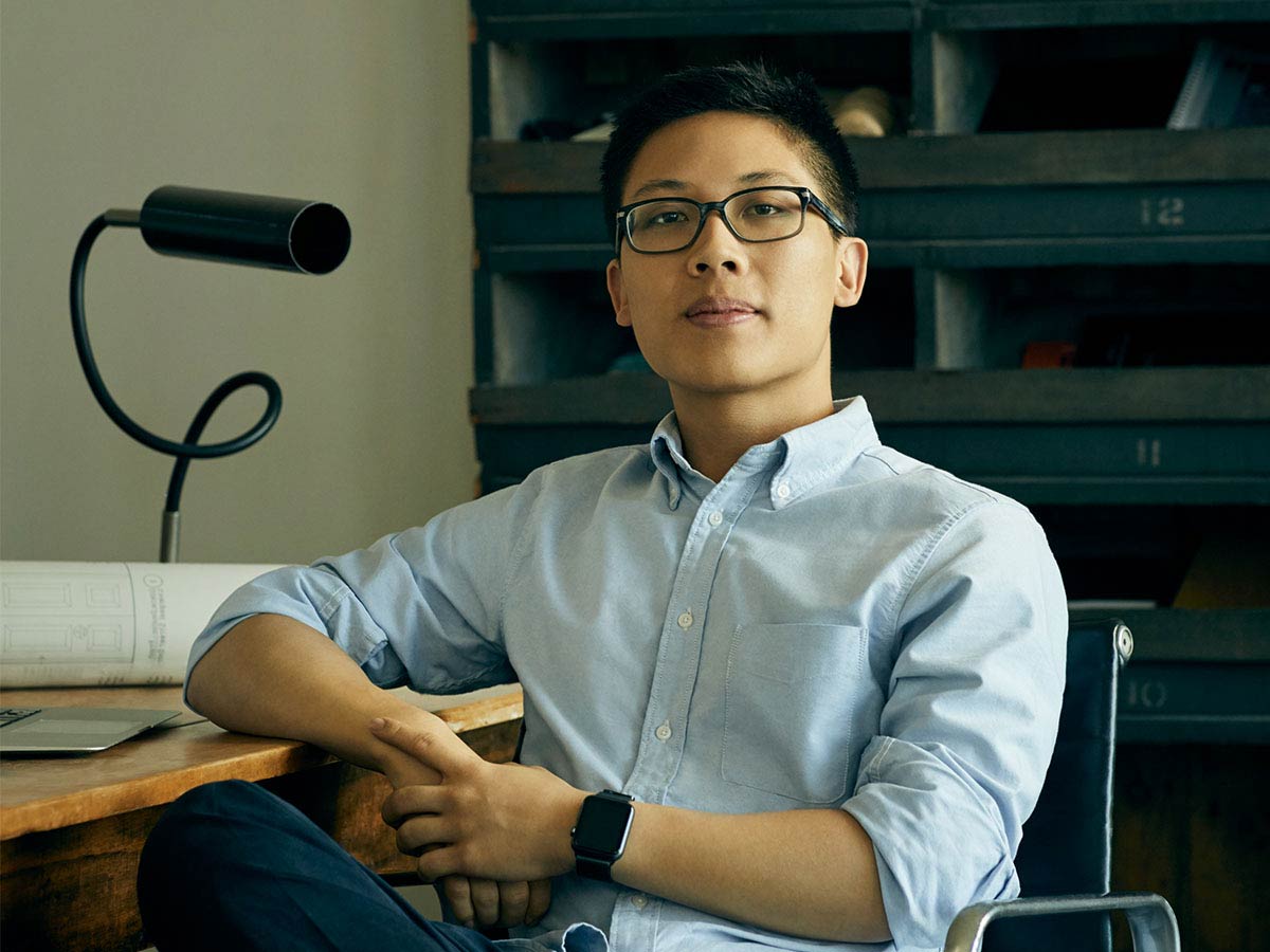 A young Asian man with glasses sitting at a desk in a casual modern office.